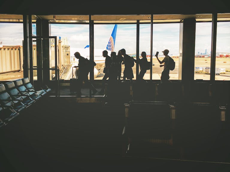 Silhouettes of travelers at an airport gate, airplane and city skyline visible through windows.