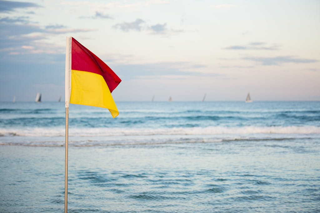 A red and yellow surf safety flag on an Australian beach overlooking a calm sea under a clear sky.