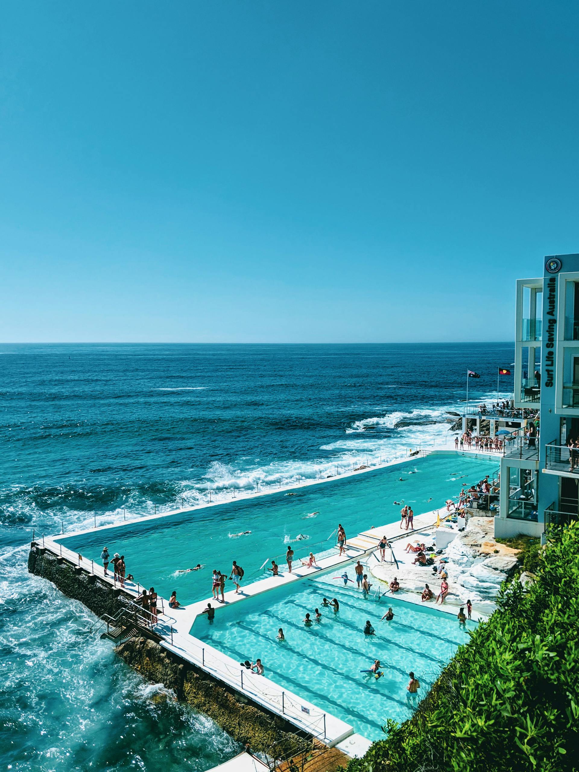 Aerial view of an oceanfront swimming pool with people enjoying a sunny day near the sea.